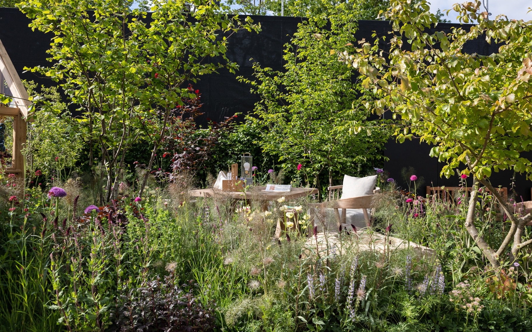 a garden with lots of plants and flowers and three trees with a wooden garden furniture in the middle with cream seat cushions