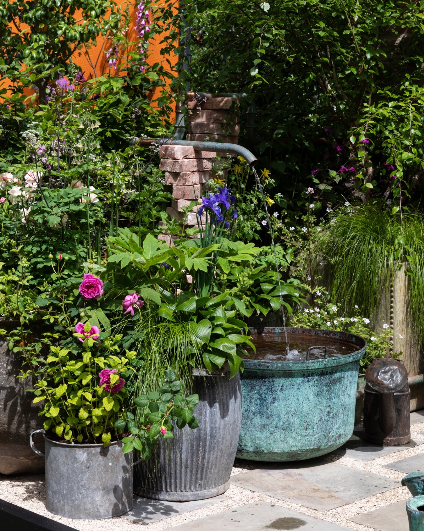 a grey patio with 3 large metal plant pots, 2 filled with greenery and pink and purple flowers and one full of water being fed by an outdoor tap