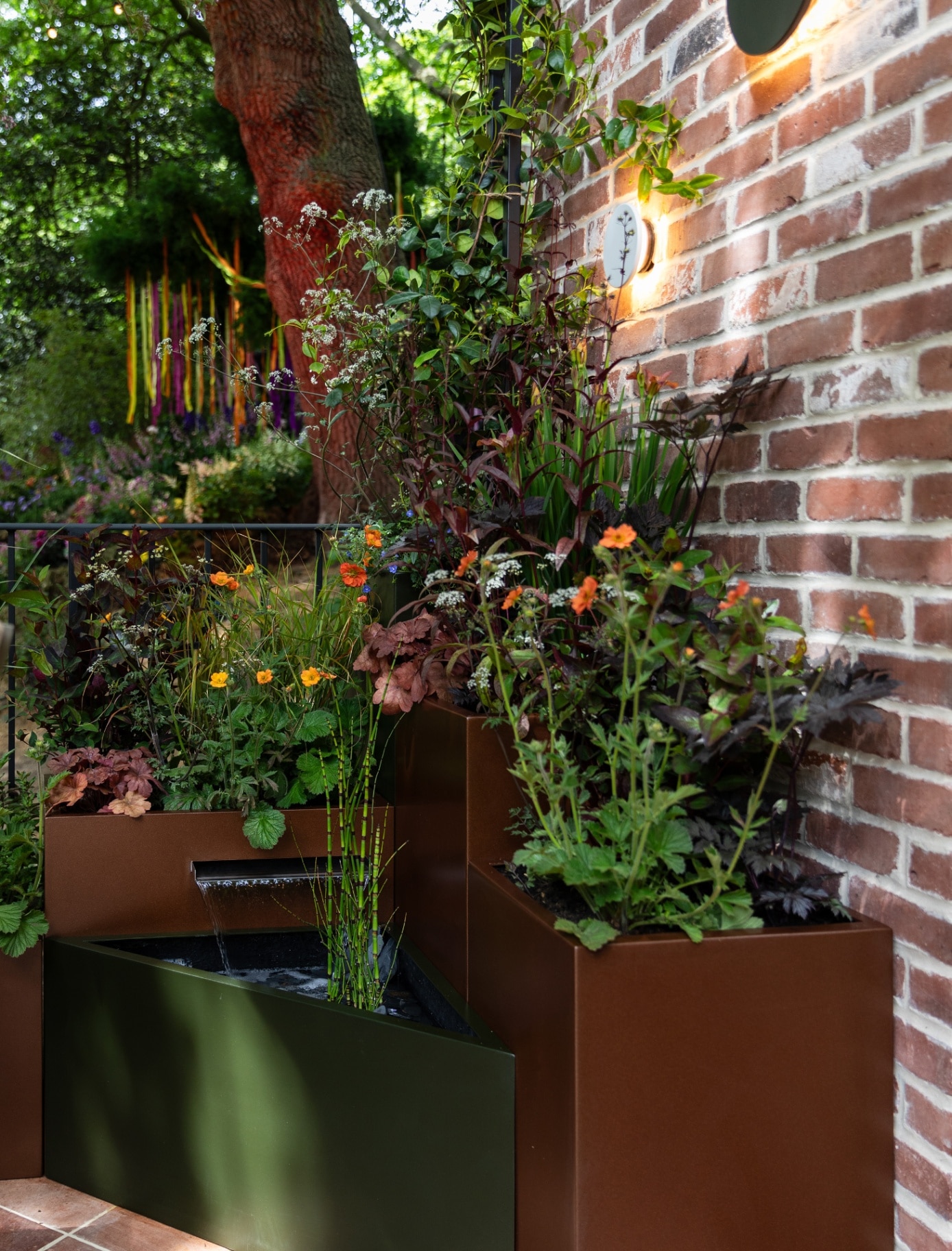 a red brick wall with two wall lights and a large brown and a large green plant pot filled with greenery and small red flowers