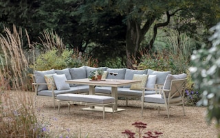 an outdoor L shape Sofa with a coffee Table and grey seat cushions on gravel in front of plants and trees