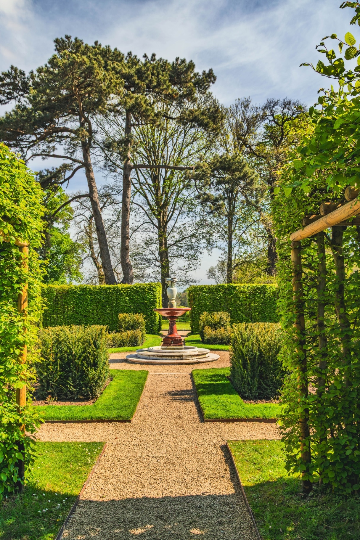 A meticulously maintained formal garden features symmetrical hedges, manicured lawns, and a decorative fountain, set against a backdrop of mature trees and a bright sky
