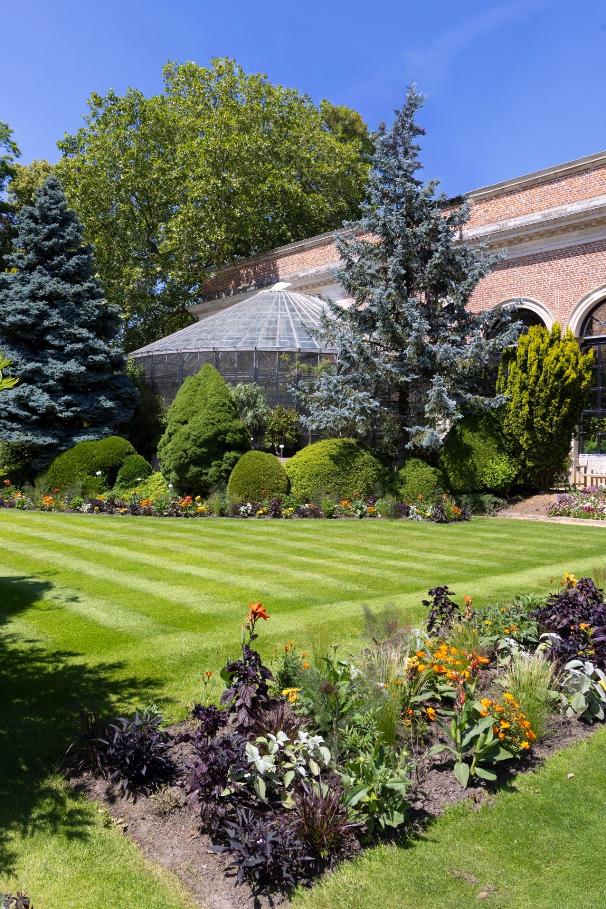 Sunlit Herbgarden (Kruidtuin) in Leuven, Belgium features a historic glass greenhouse, striped formal lawn, trimmed topiary and vibrant flower beds, offering a peaceful, colourful urban botanical escape for visitors.